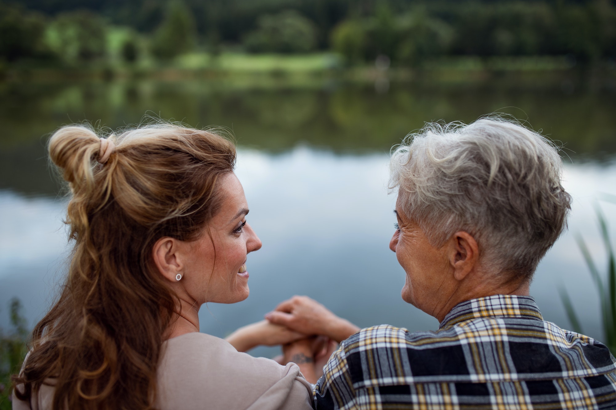 Rear view of happy senior mother holding hands with adult daughter when sitting by lake outdoors in